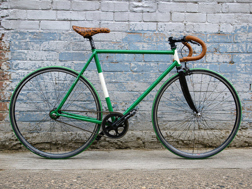 VERDE classic green fixed-gear bicycle with white frame accents, brown leather-style saddle and handlebars, photographed against a weathered brick wall. Product of Triflex Creative Studio.